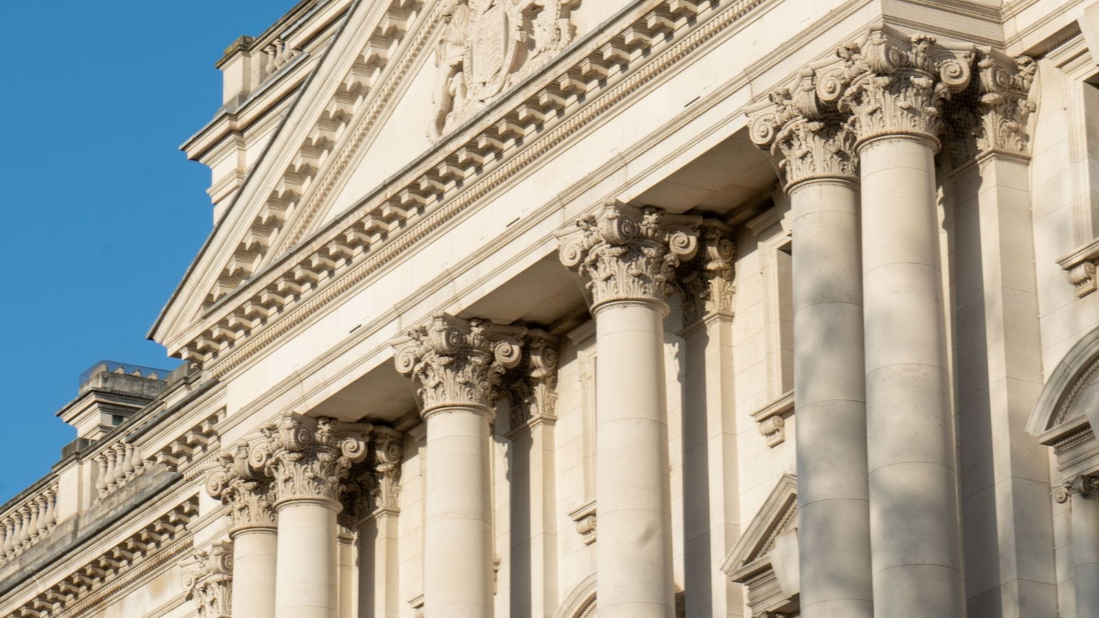 The colonnaded stone facade and royal coat of arms of a Whitehall government building against a clear blue sky