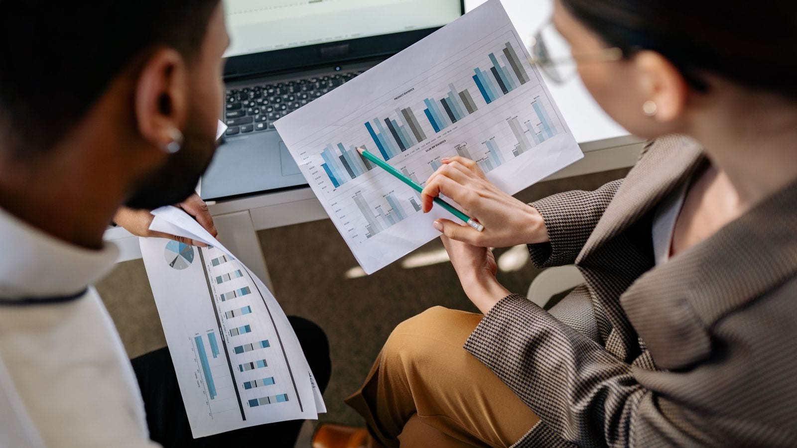 Two colleagues holding printed reports of bar charts side by side, one pointing at a row with a pencil